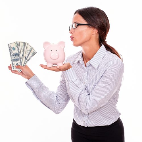 Businesswoman holding pink porcelain piggy bank in one hand and money with the other hand while blowing a kiss at the piggy bank and wearing her hair back in a button down shirt on a white background